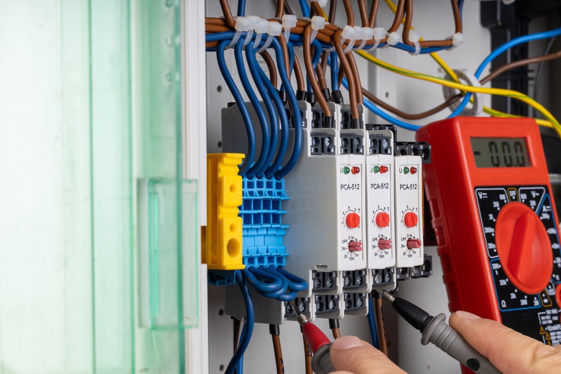 Electrician testing circuits in a control panel with a multimeter, wires are blue, orange, and brown.