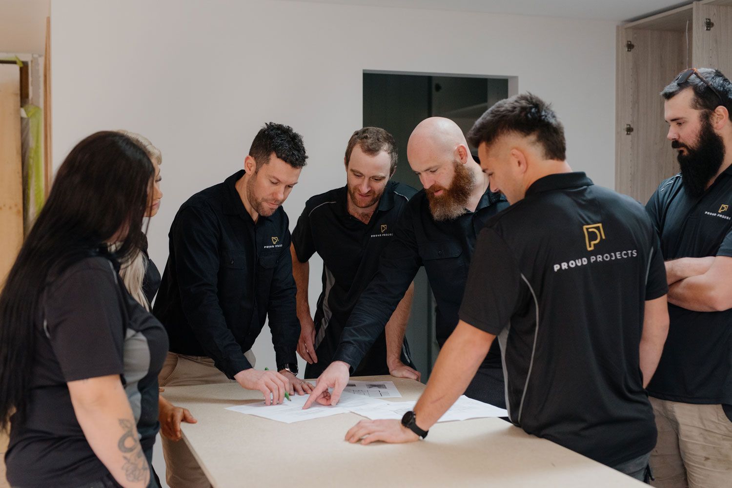 Group of people in black shirts looking at a blueprint on a table indoors.