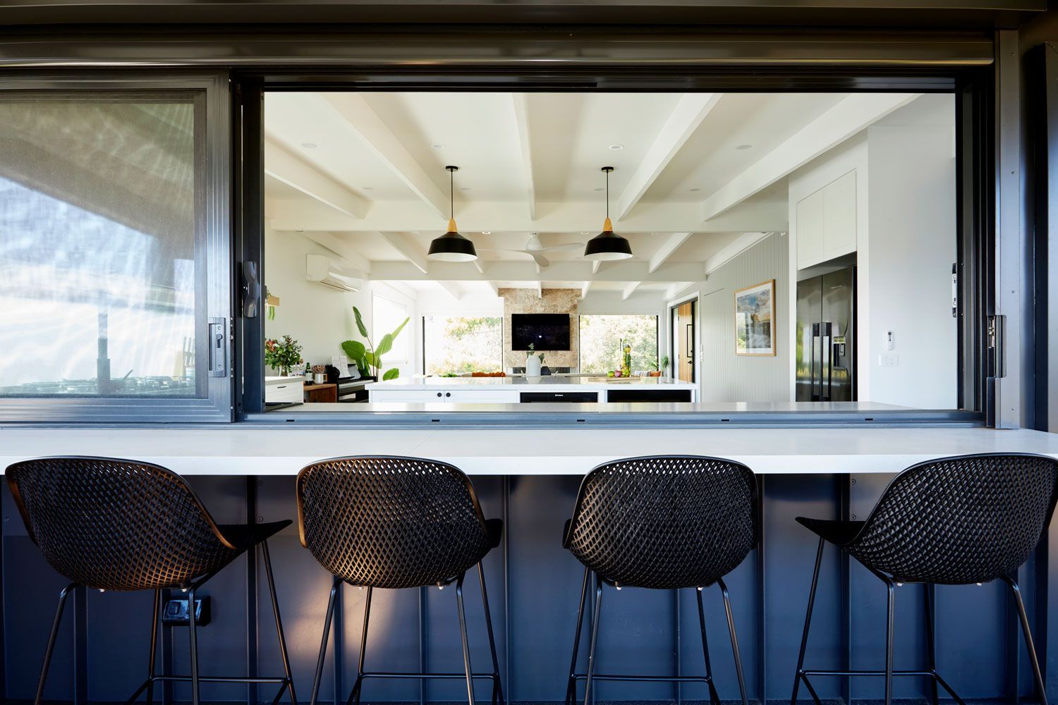 Four black wicker bar stools sit at a window overlooking a white kitchen with pendant lights.