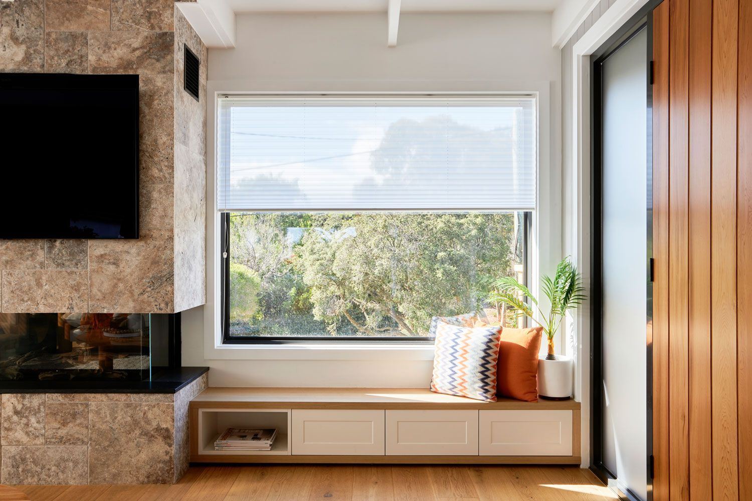 Living room window with a window seat, built-in storage, and two throw pillows. Sunlight streams through the trees.