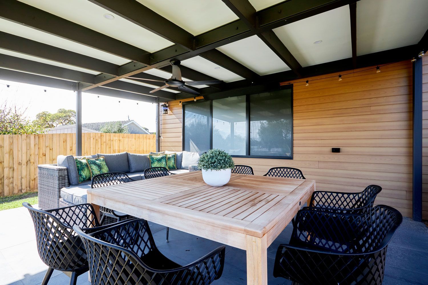 Outdoor patio with wooden table, black chairs, and a seating area with cushions and plants.