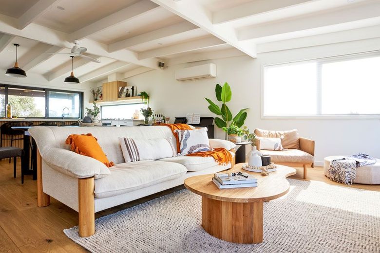Bright living room with white sofa, wood coffee table, and plants; kitchen in the background.
