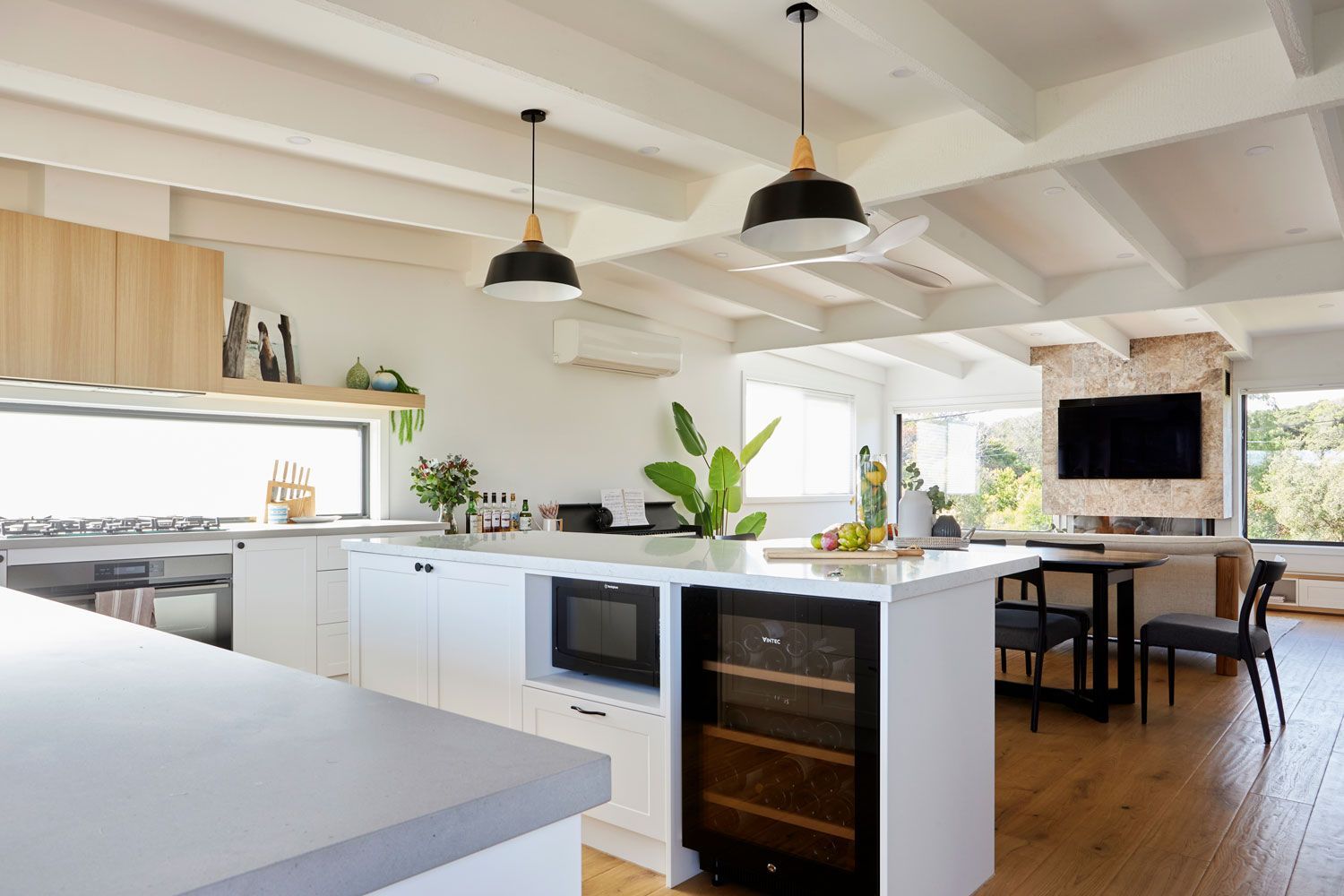 Modern white kitchen with island, wooden floors, black pendant lights, and large windows.