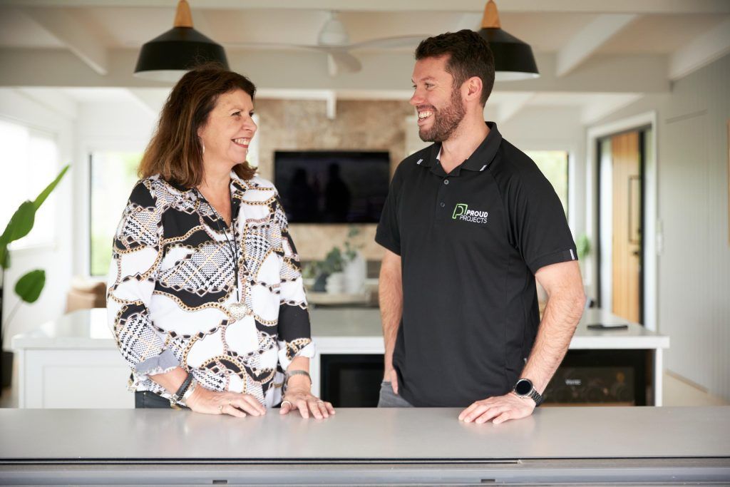 Woman and man smiling at each other in a kitchen. The man wears a black polo, the woman a patterned blouse.