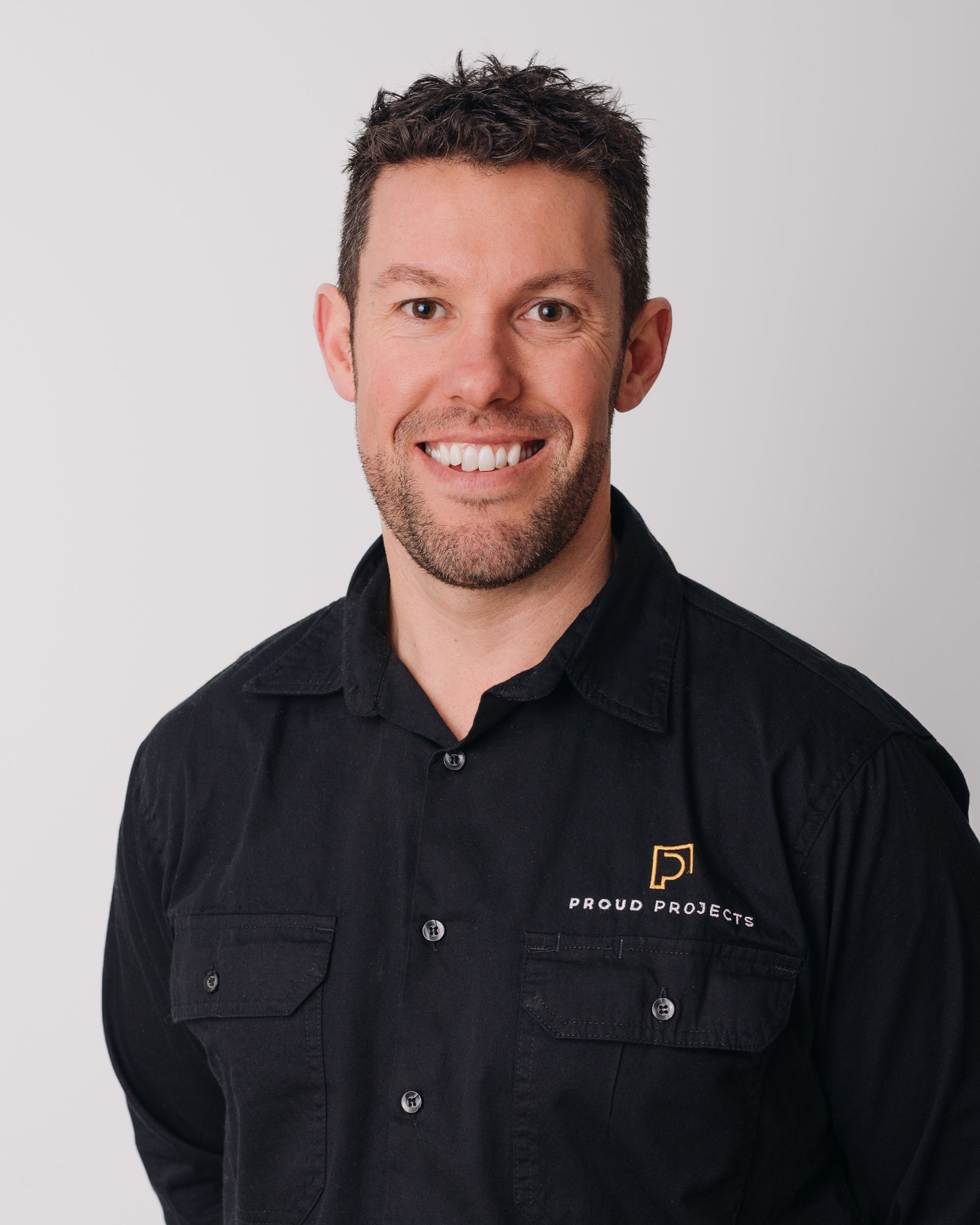 Man smiling, wearing a black shirt, logo on chest, against a white background.