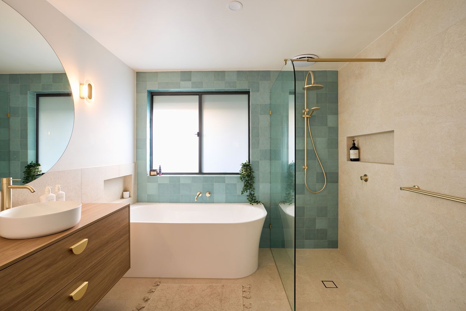 Modern bathroom with teal tile, freestanding tub, wooden vanity, and gold fixtures.