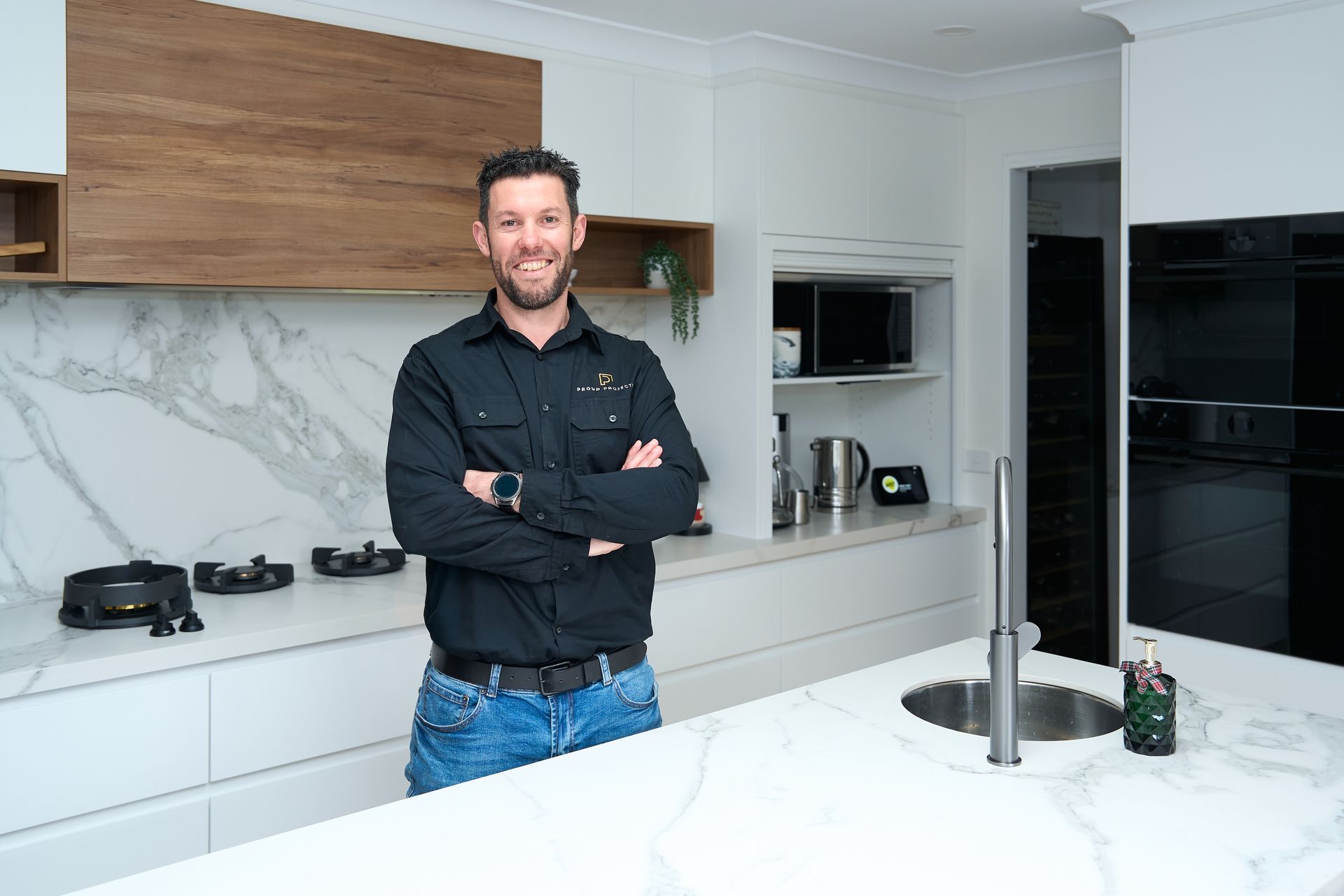 Man with arms crossed smiles in a white and wood kitchen, standing near an island with a sink.