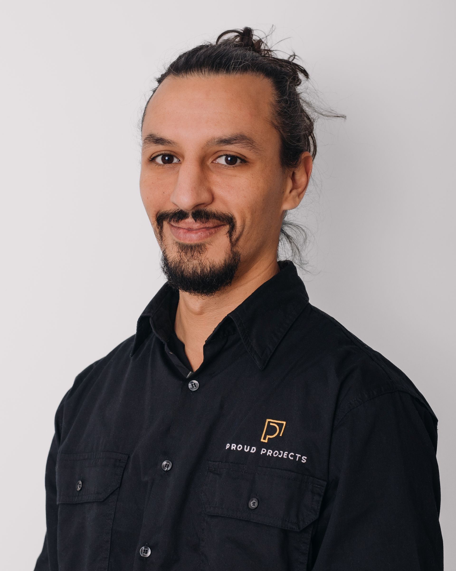 Man in black shirt with logo, hair in a bun, smiling, on a white background.