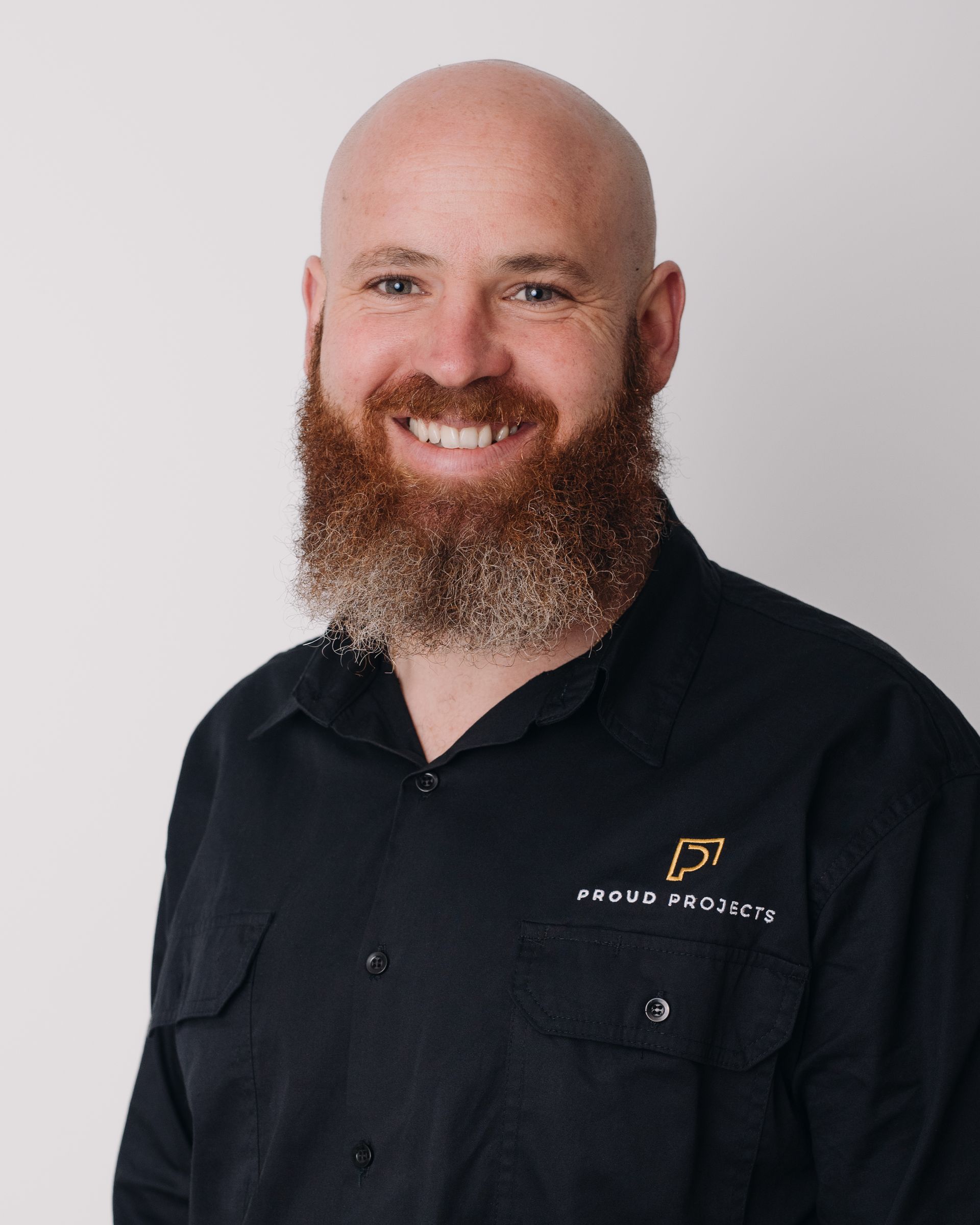Bald man with a red beard wearing a black shirt with logo, smiling against a white background.