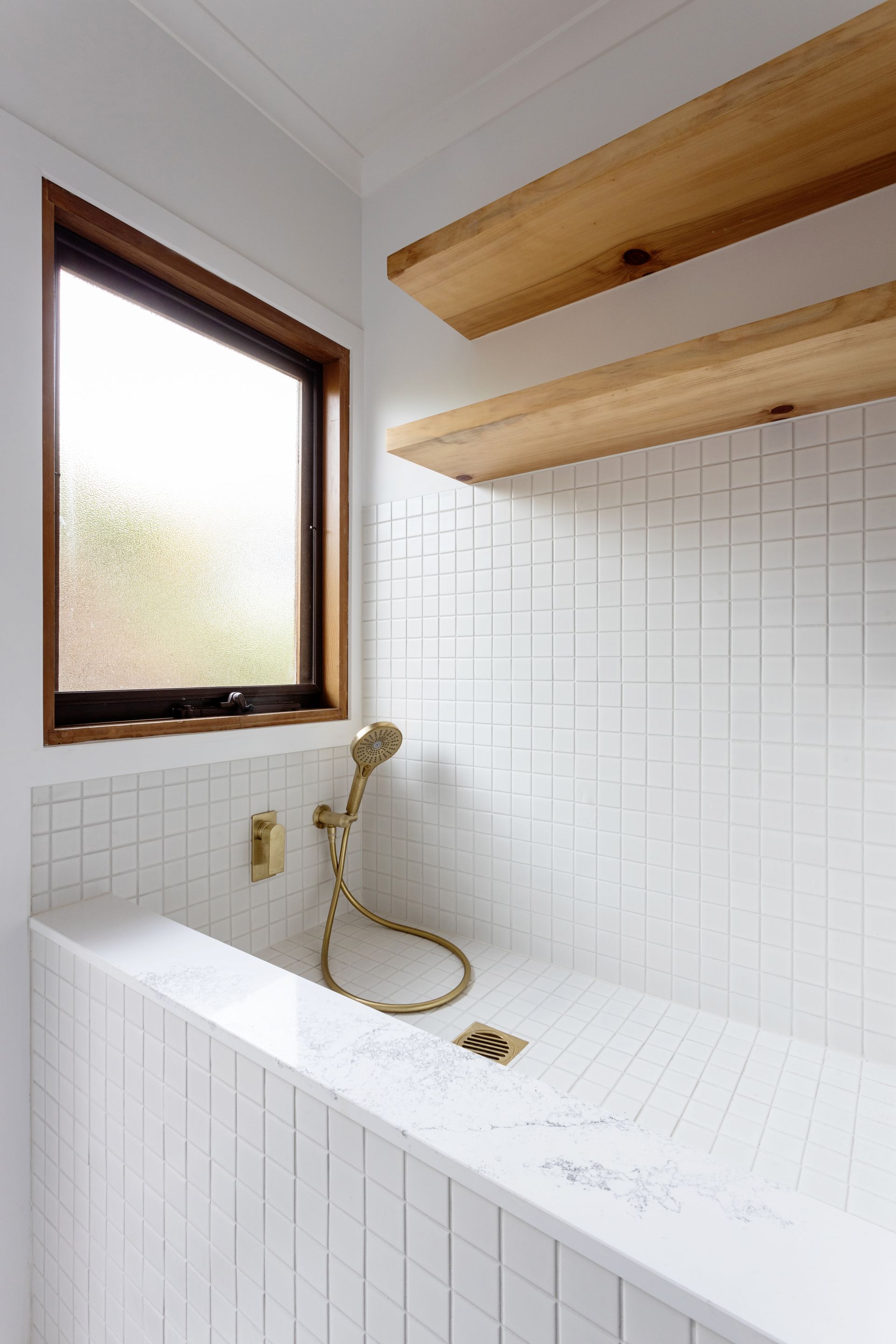 Modern white tiled bathroom with built-in tub, golden fixtures, wooden window frame, and shelves.