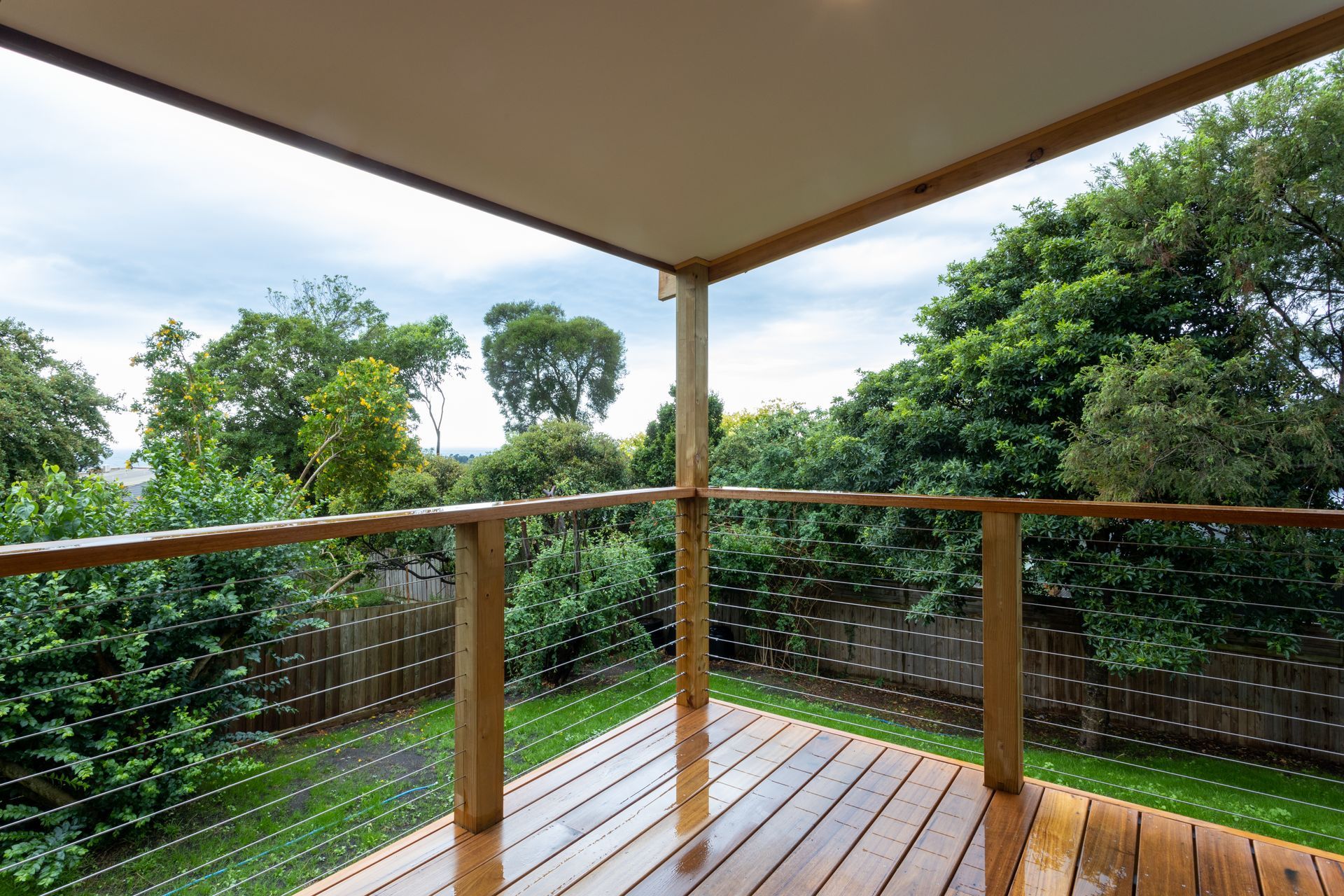 Wooden deck with wire railings overlooking a lush green yard and trees. Overcast sky.