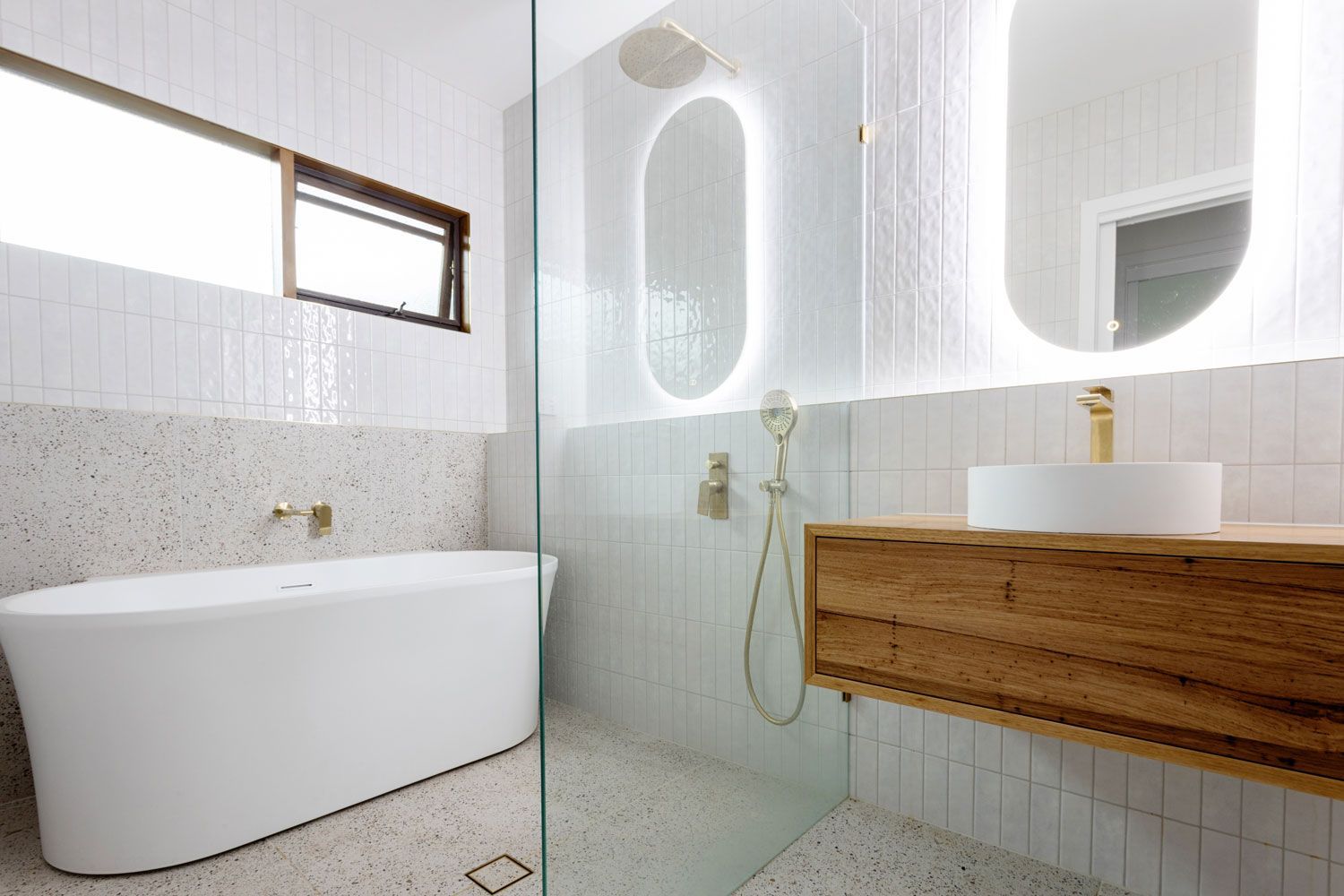 Modern bathroom with white tub, wood vanity, and gold fixtures. White tile walls and oval mirror with LED lighting.