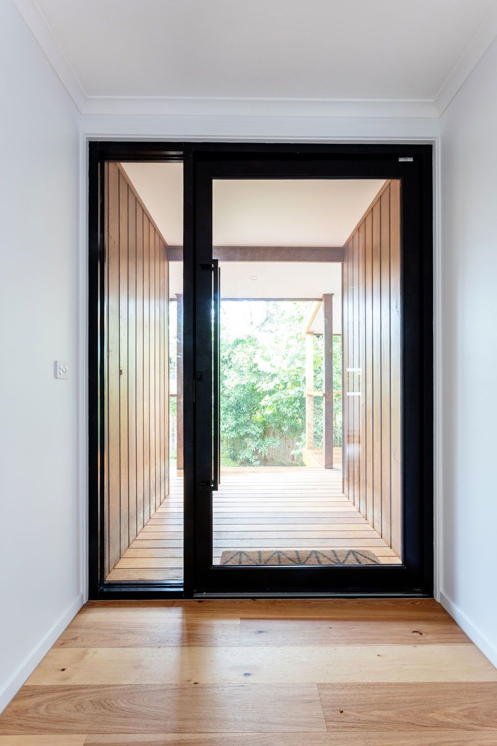 Black-framed glass door with sidelight opens onto a wood-lined walkway leading to a green outdoor space.