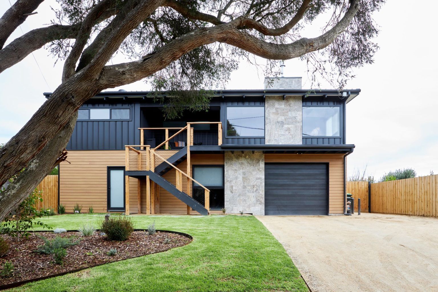Modern two-story house with black and tan siding, stone facade, a garage, and an outdoor staircase under a large tree.
