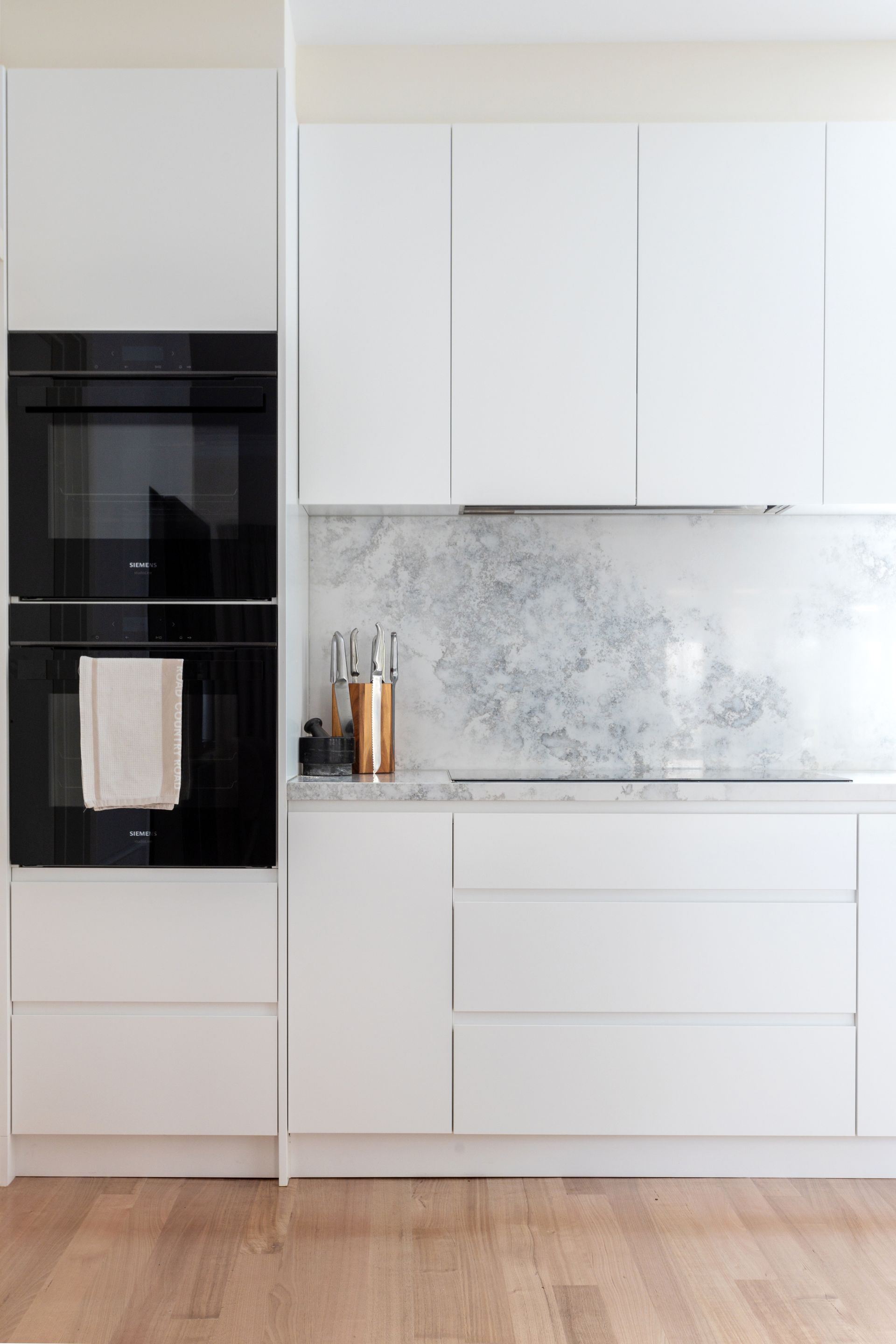 Modern white kitchen with built-in black oven, white cabinets, marble backsplash, and wooden floor.