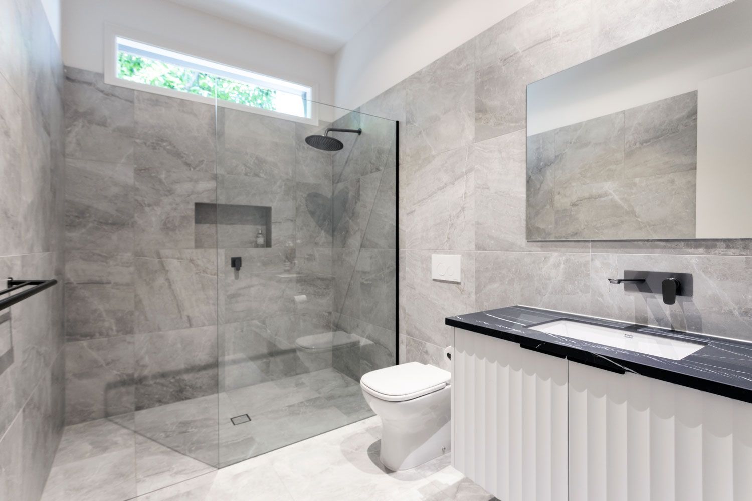 Modern bathroom with gray marble-look tile, glass shower, black fixtures, white vanity, and rectangular mirror.