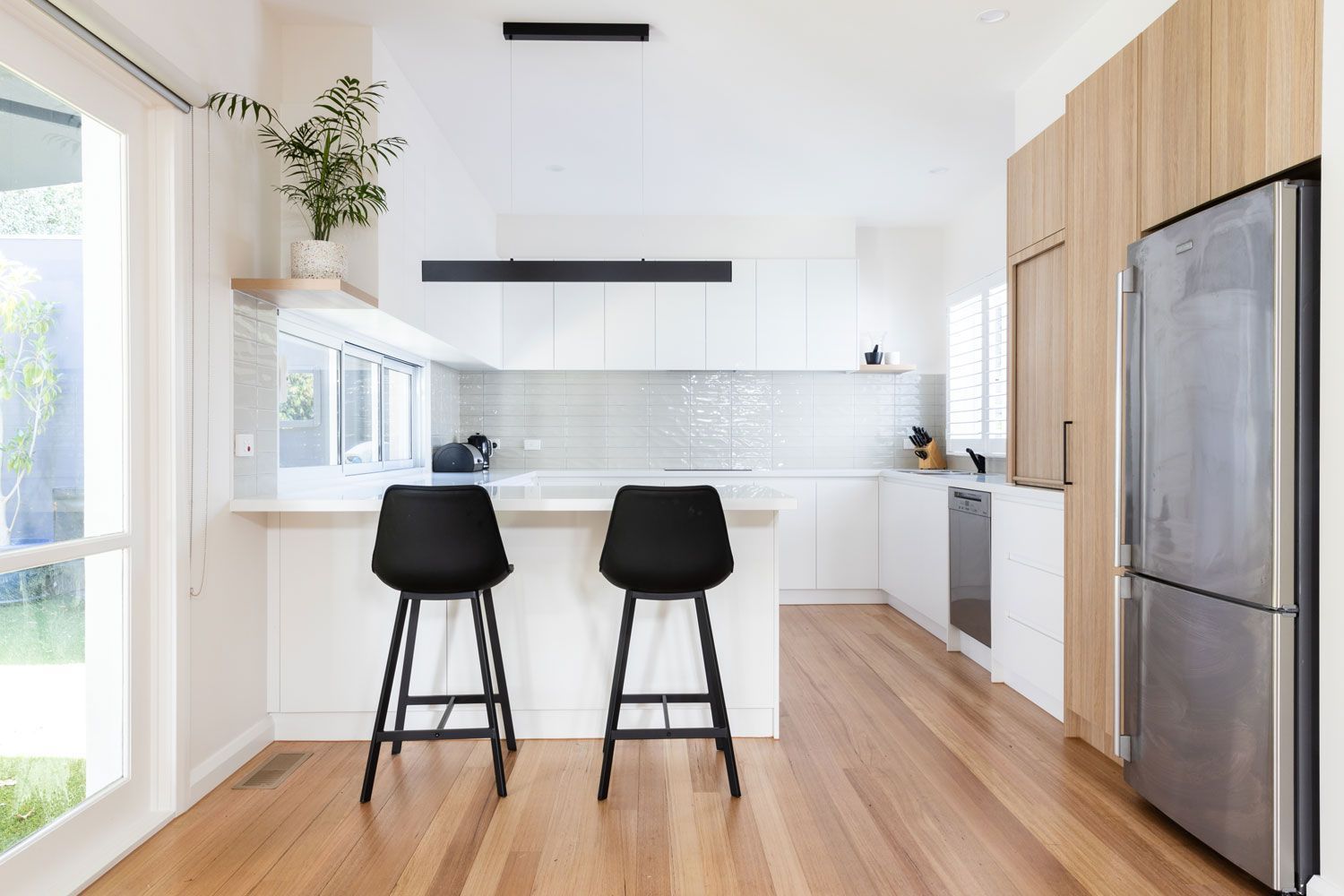 Modern kitchen with white cabinets, stainless steel refrigerator, wooden floors, and two black bar stools.