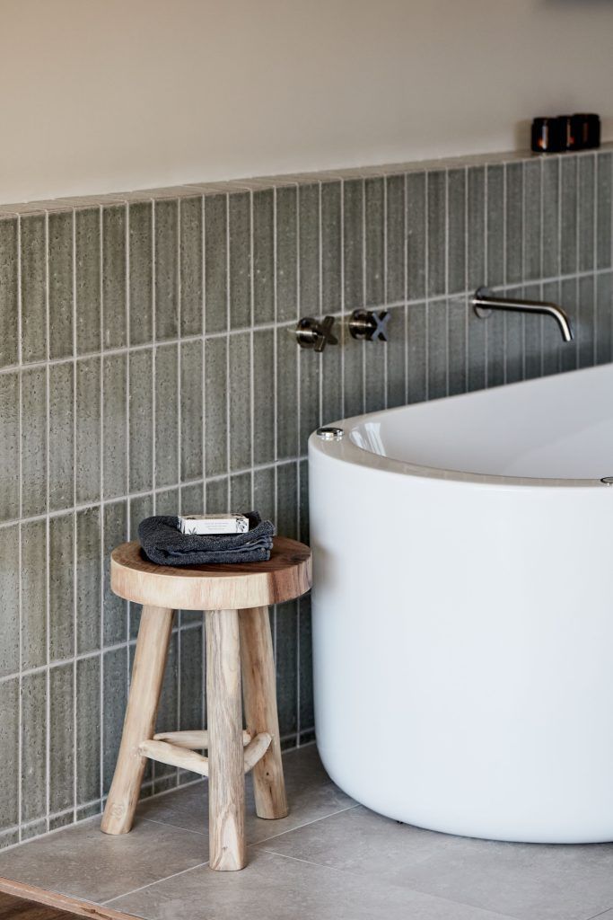 Bathroom with a white curved bathtub, gray vertical tiles, a wooden stool, and silver faucet.