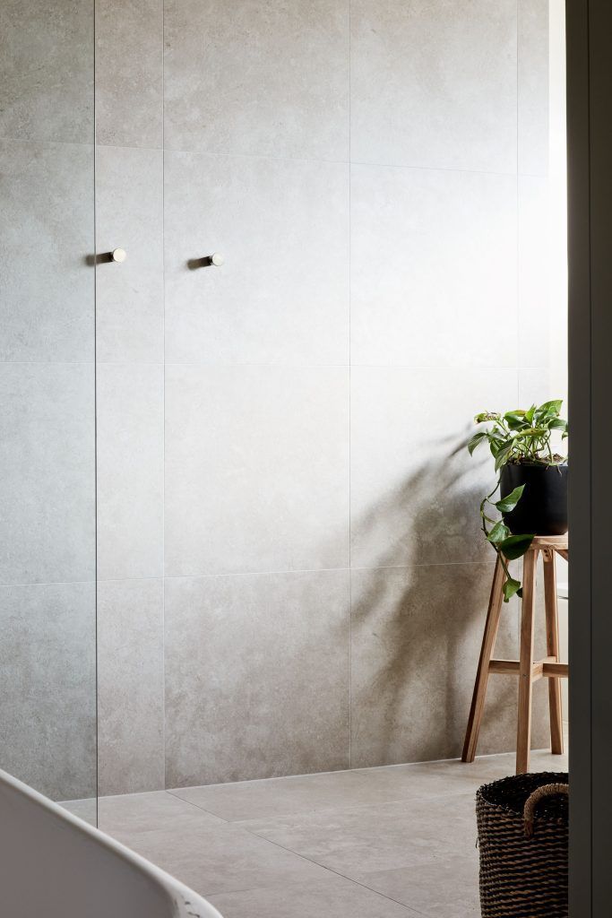 Light gray tiled bathroom, plant on wooden stool, gold towel hooks.