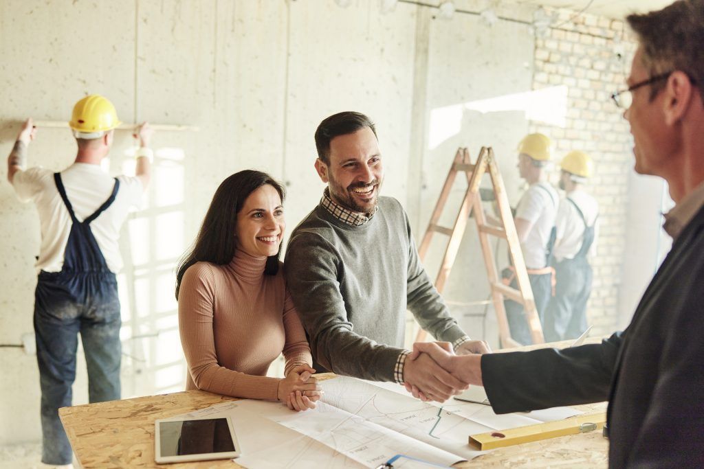 Couple shaking hands with a professional at a construction site; blueprints, workers in background.