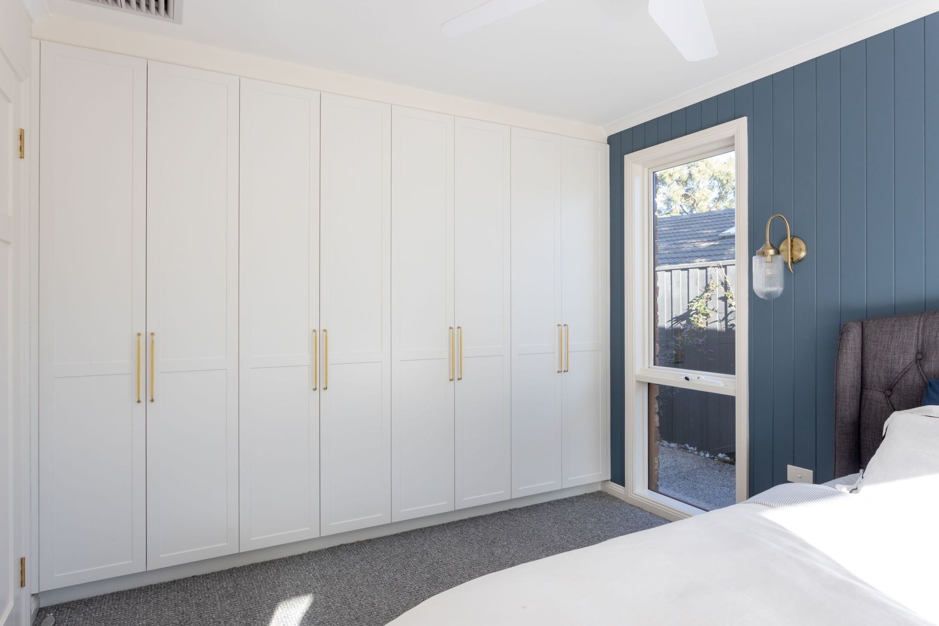 White closet doors with gold handles in a bedroom; blue accent wall with a window and a bed.