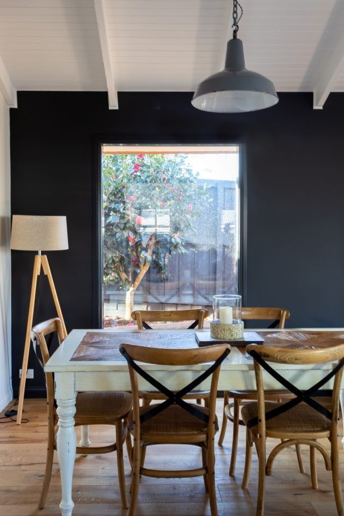Dining room with black wall, white table, wooden chairs, and a window overlooking a tree.