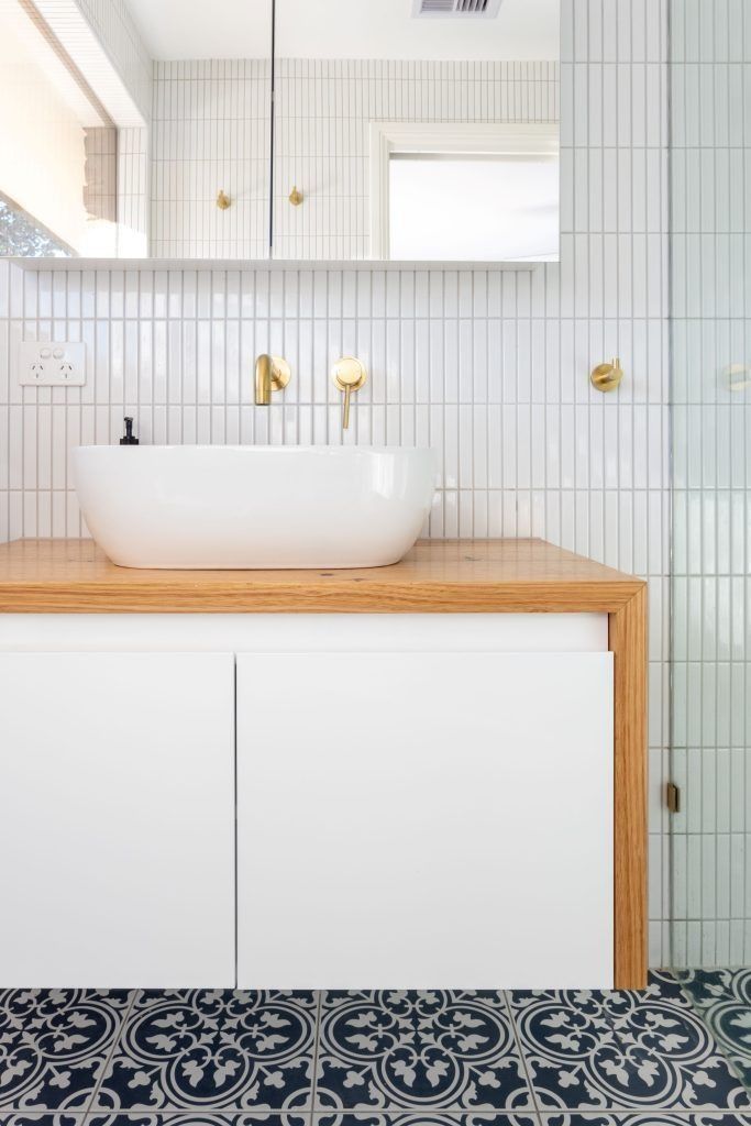 White bathroom with a wood vanity, gold fixtures, patterned floor tiles, and vertical white wall tiles.