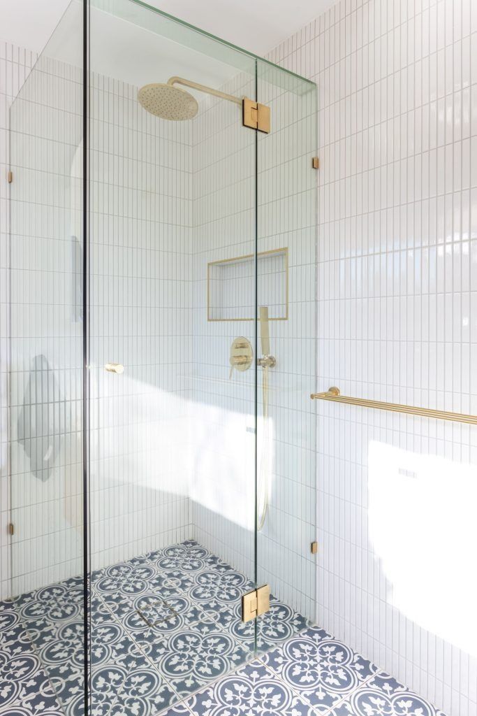Glass shower with gold fixtures, white tiled walls, and patterned blue and white floor tiles.