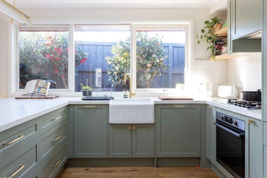 Sage green kitchen with white countertops and a large window overlooking a backyard.