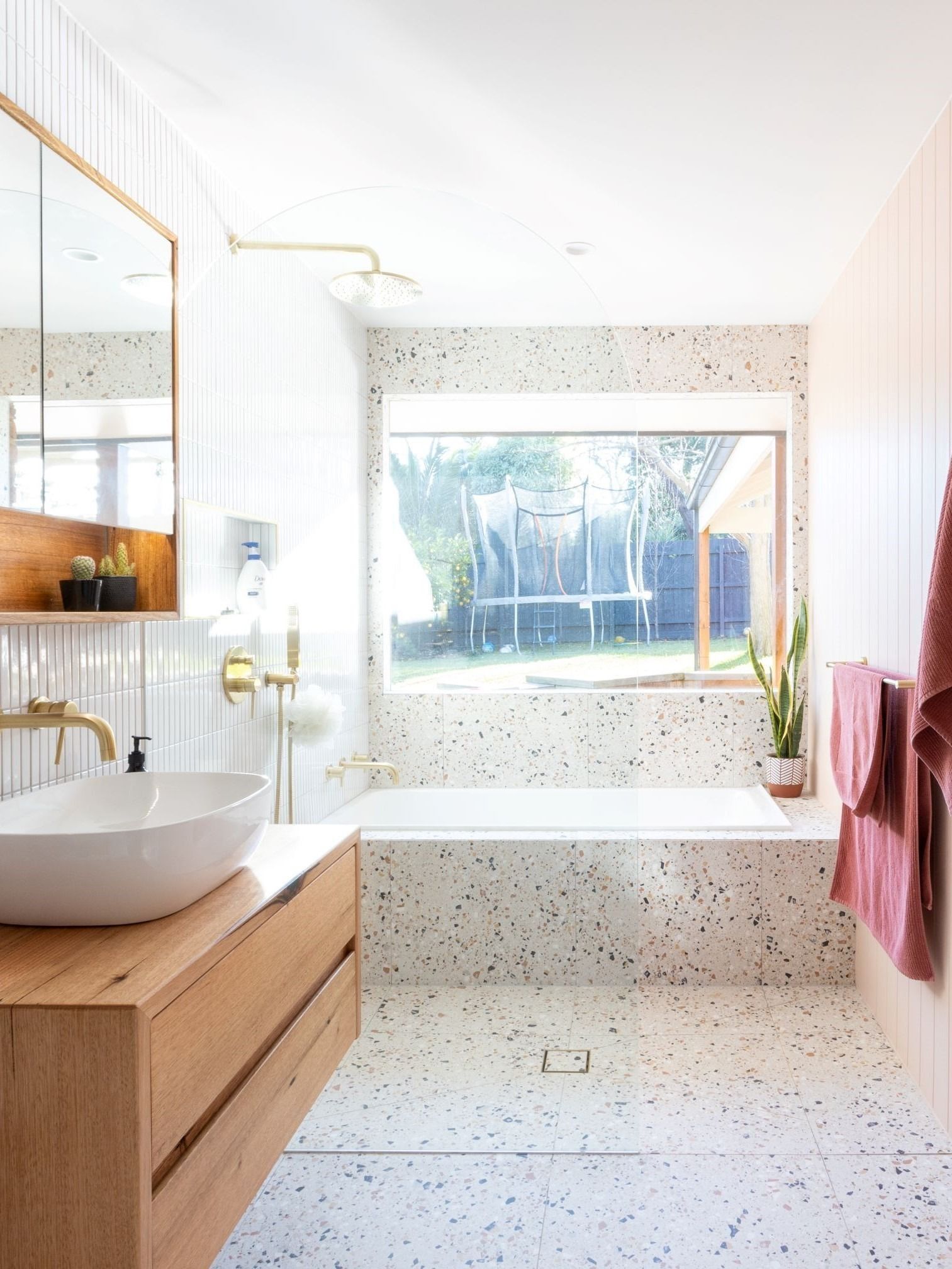 Bathroom with terrazzo floor, wood vanity, bathtub under a window, and brass fixtures.
