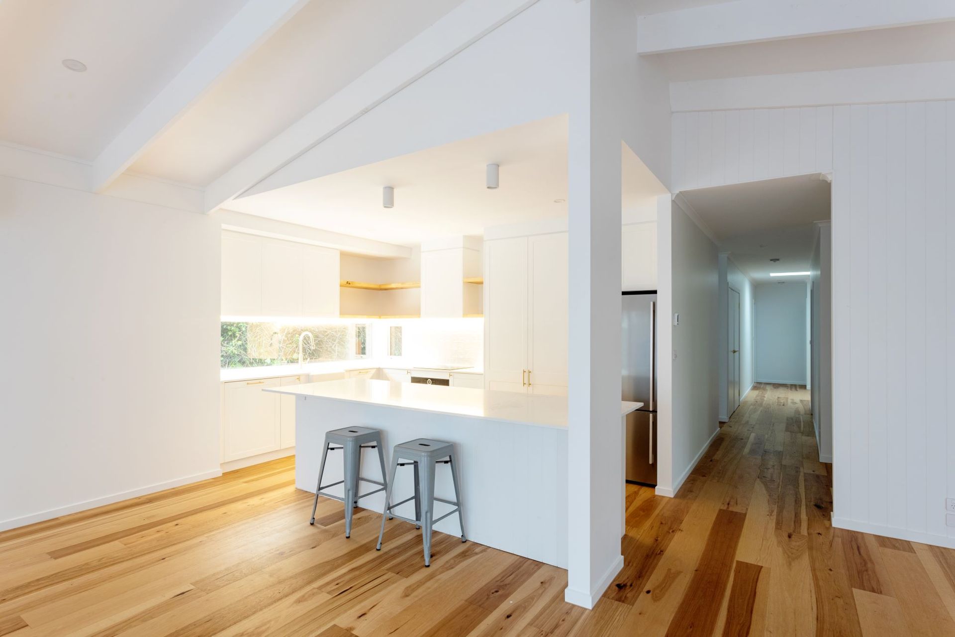 Bright, modern interior with hardwood floors. Kitchen island with stools, white walls, and a hallway.