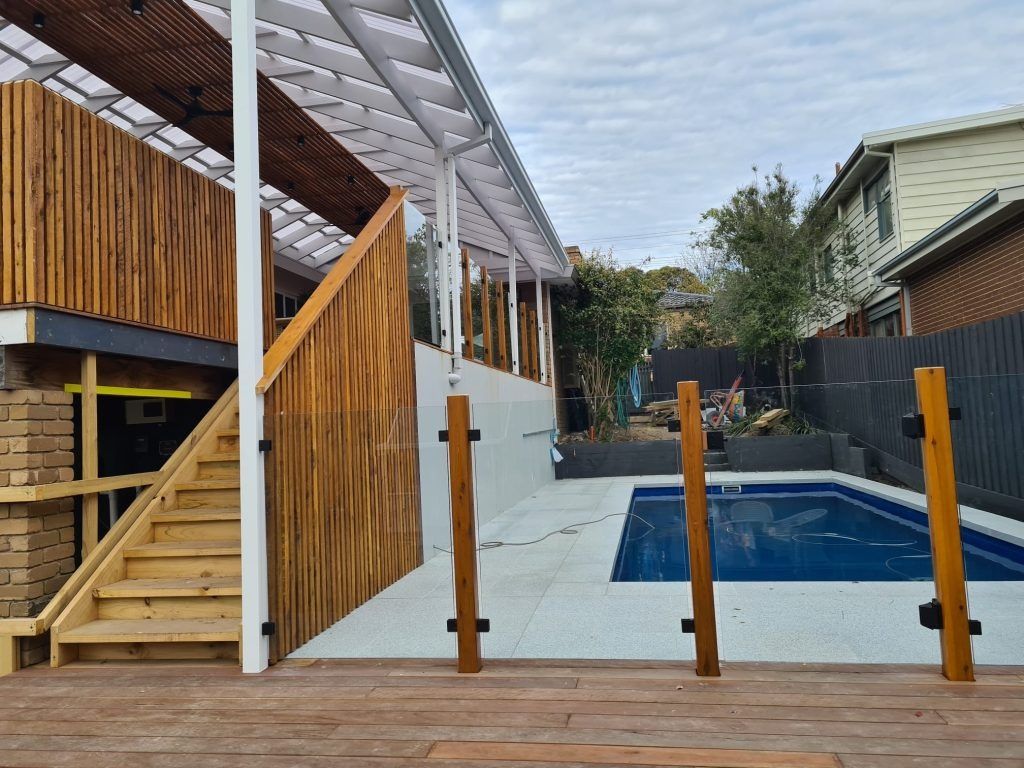 Wooden deck with stairs leading to a poolside area with a blue pool. Buildings surround the pool.