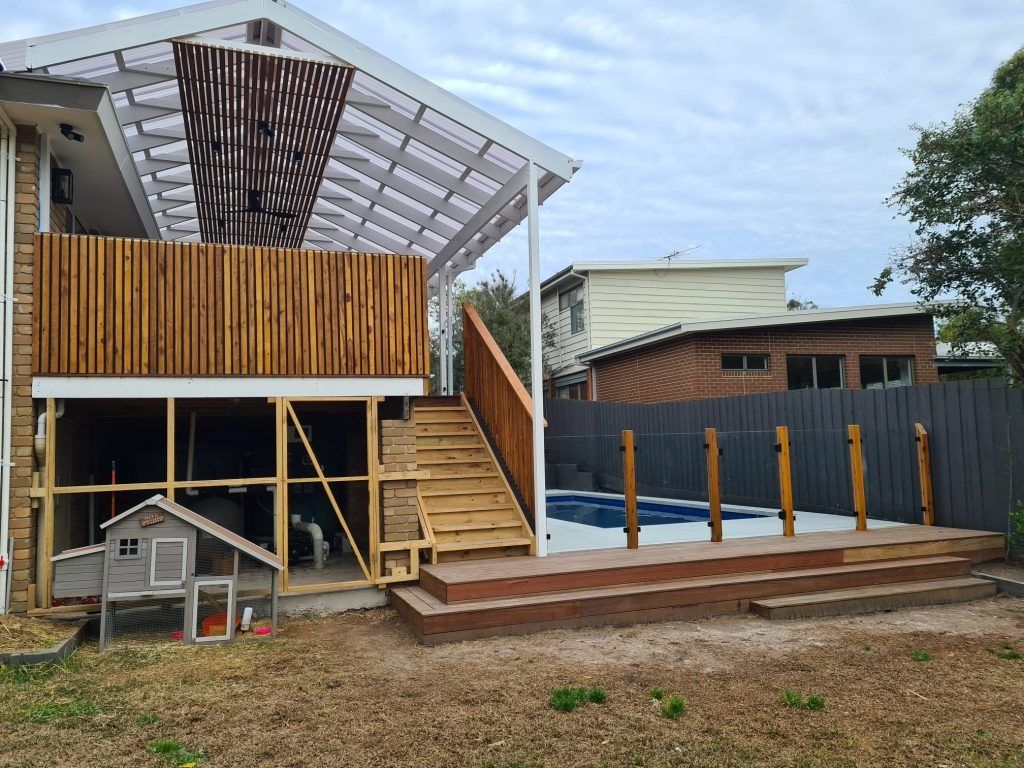 Backyard with a wooden deck, stairs, and a pool. A chicken coop sits nearby. Overcast sky.