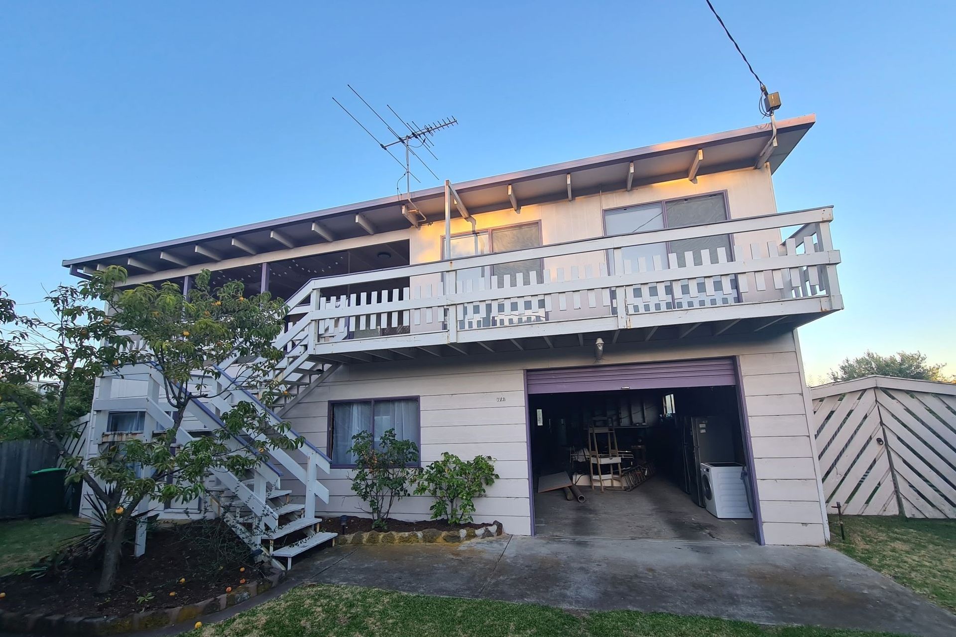 Two-story beach house with open garage. White balcony, pink siding, grass lawn, blue sky.