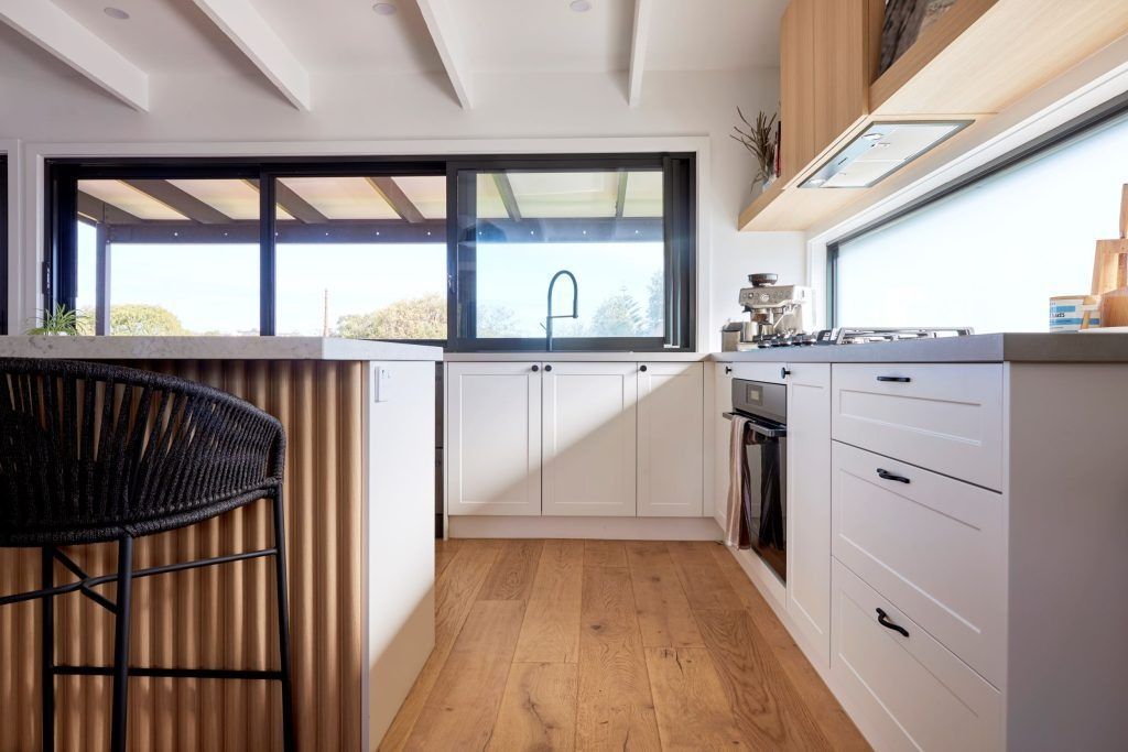 Modern kitchen with white cabinets, wooden floor, and corrugated metal island. Black stool in foreground.