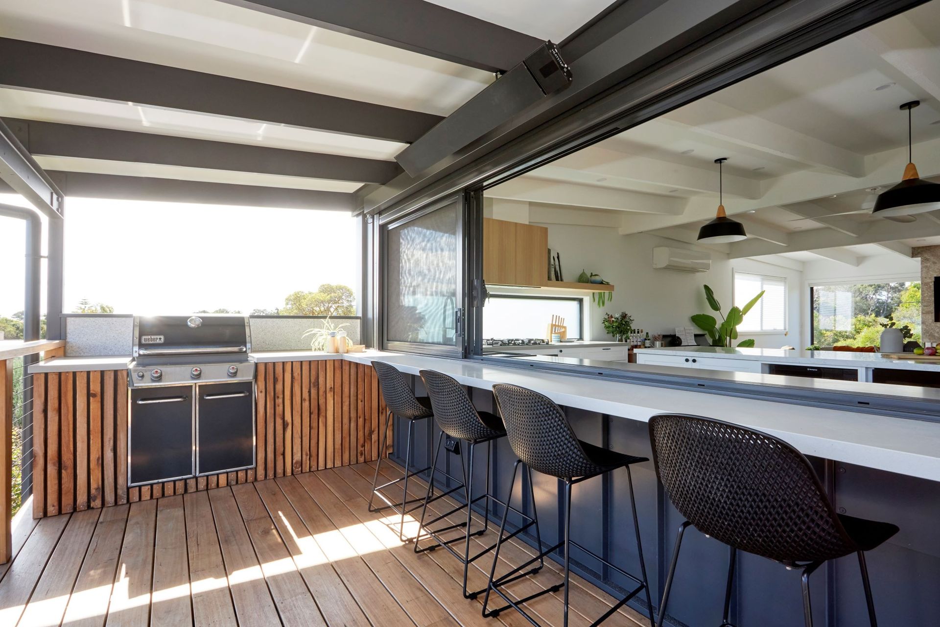 Outdoor kitchen with grill, bar, and seating, overlooking the interior. Wooden accents, white countertops.