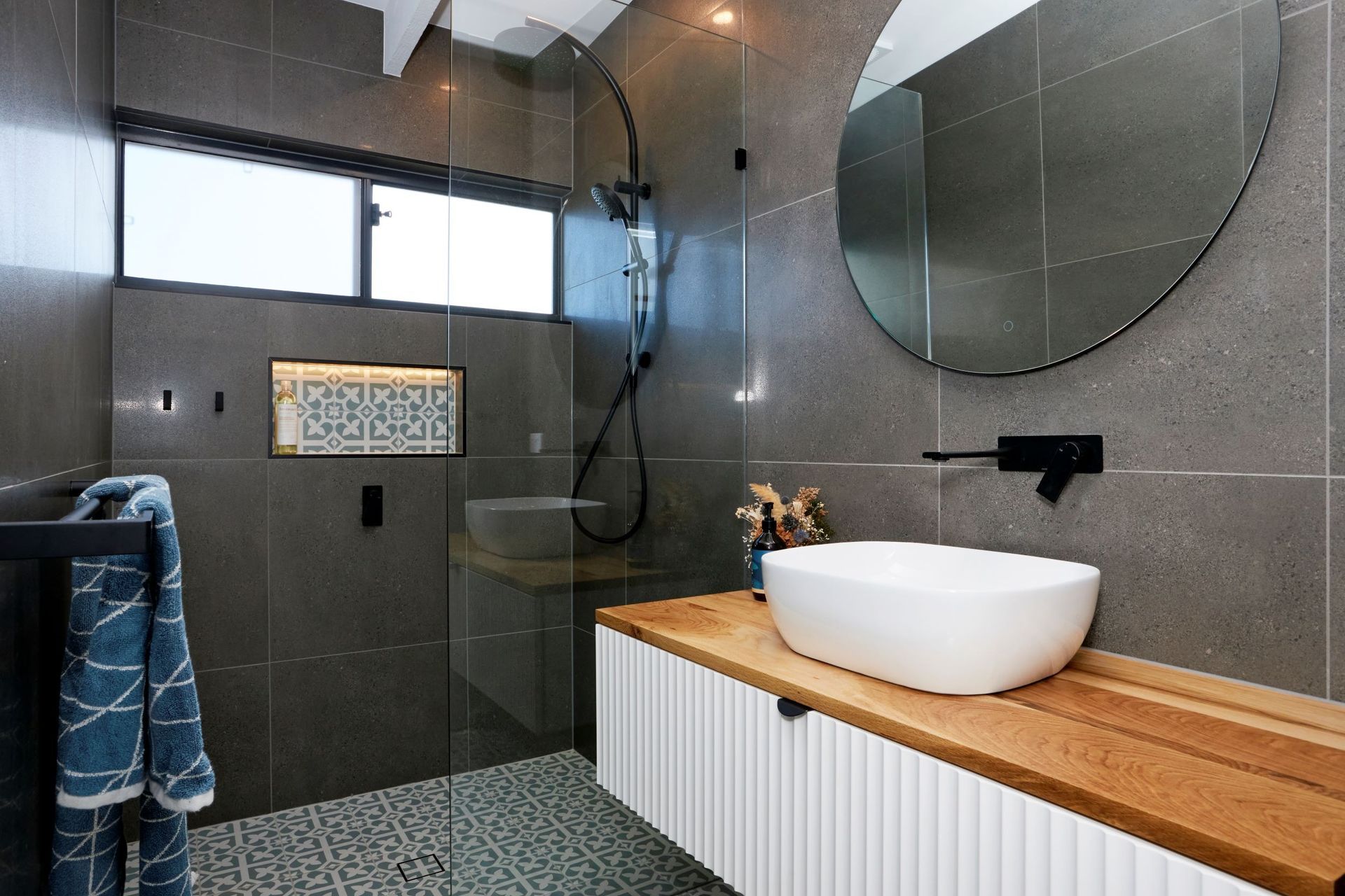 Modern bathroom with a glass shower, floating vanity, and round mirror; gray tiles and wooden countertop.