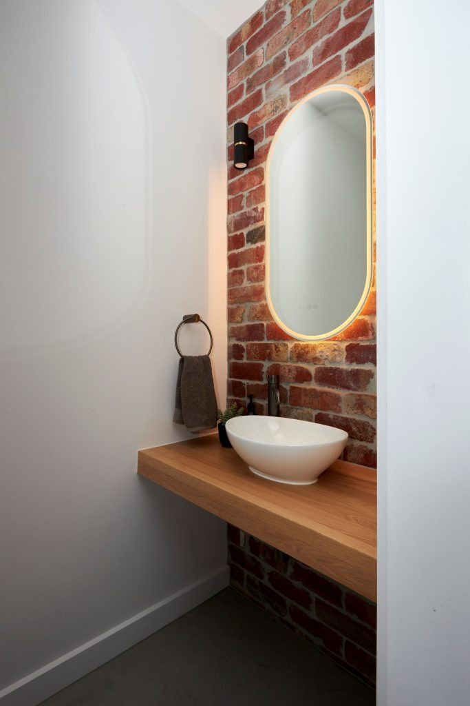 Powder room with exposed brick wall, floating wood shelf, oval mirror with warm lighting, and white vessel sink.