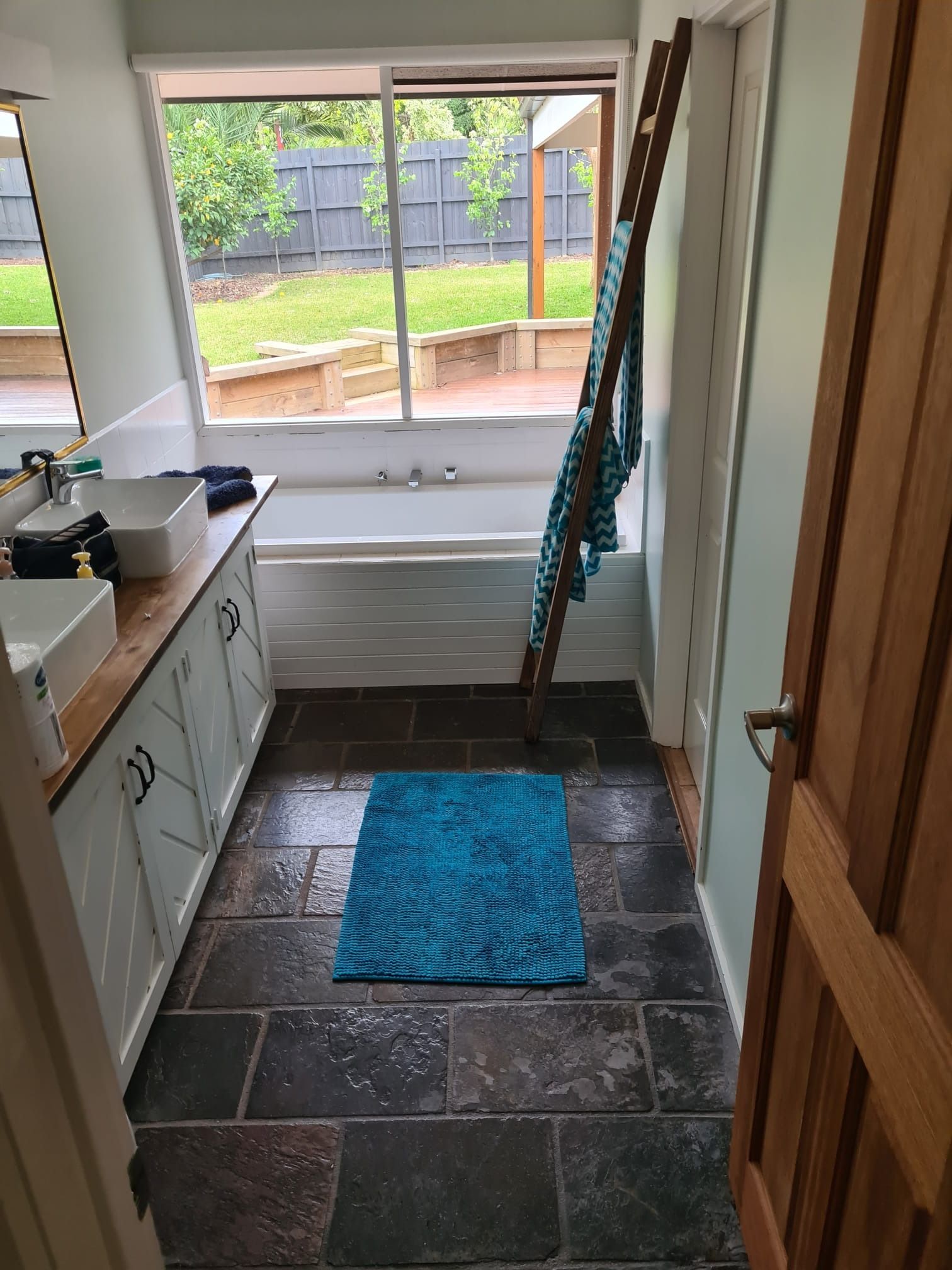 Bathroom with a bathtub, two sinks, and a blue bath mat. A towel ladder leans against the wall.