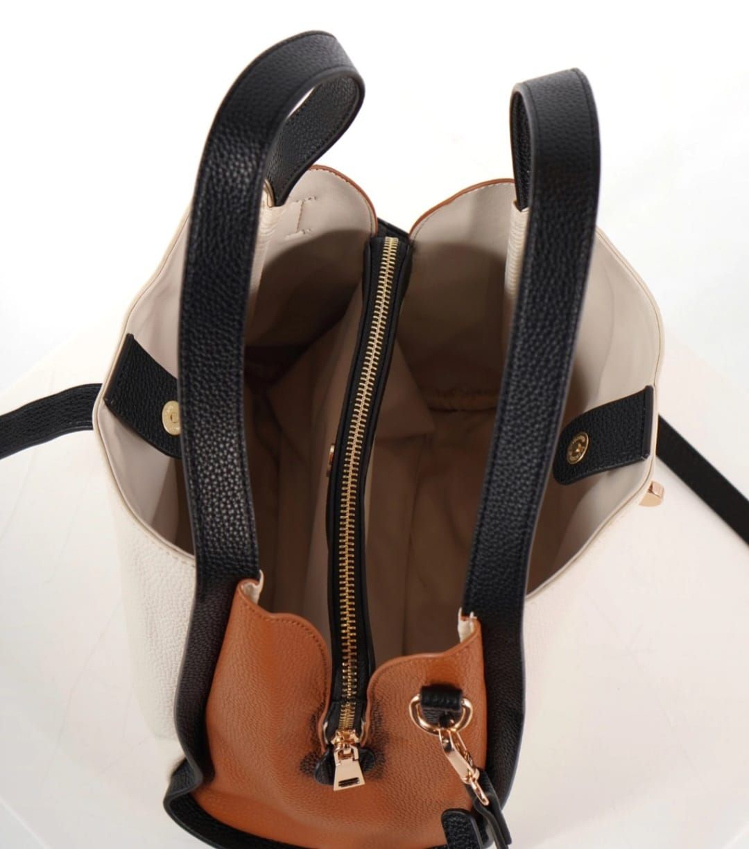 Bolso color crema con ribete, asas y correa en negro. Tiene cierre de hebilla y reposa sobre una superficie blanca.