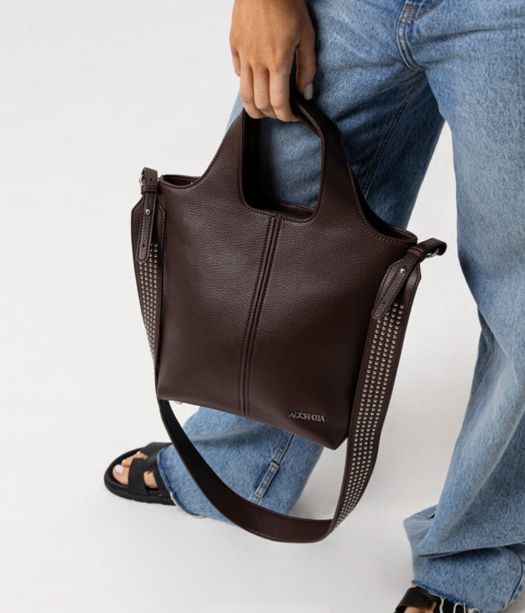 Bolso de mano de piel marrón con bandolera con tachuelas y asa corta.