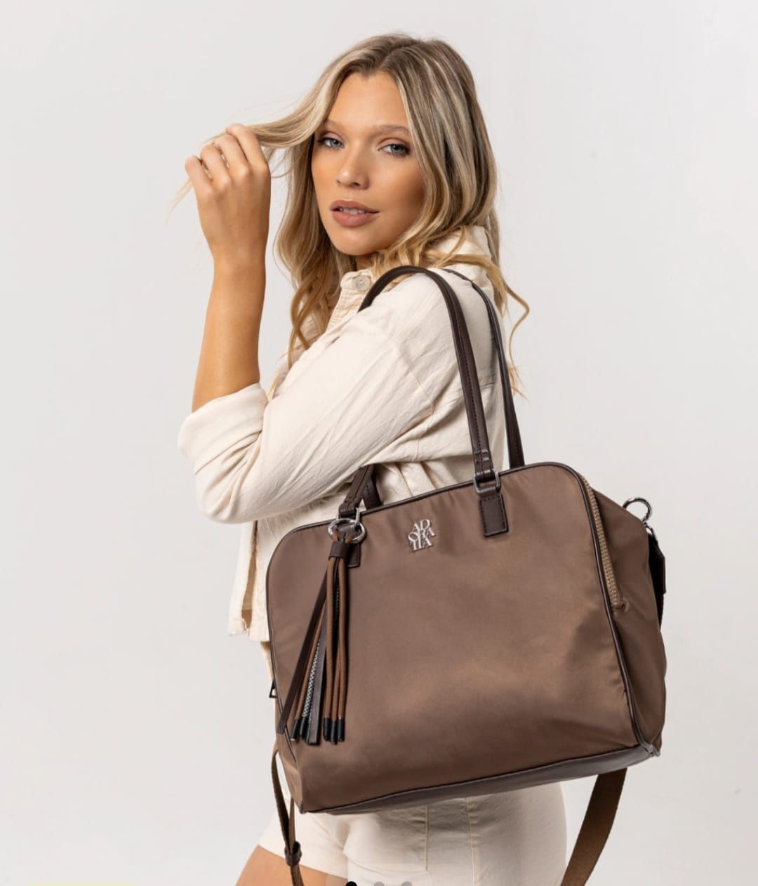 Mujer sosteniendo un bolso marrón, vistiendo una chaqueta color crema, mirando hacia la cámara.