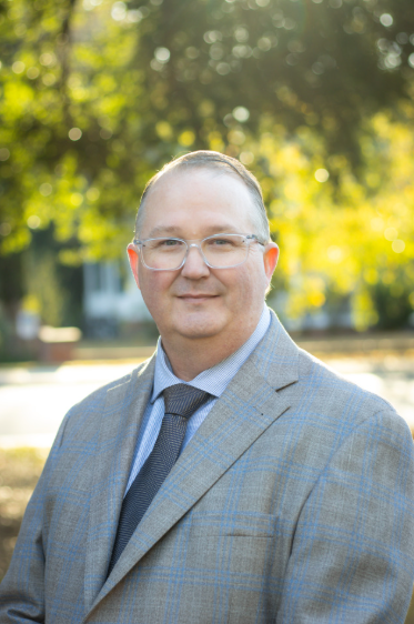 Man in a gray suit and glasses, smiling, outdoors in front of trees.
