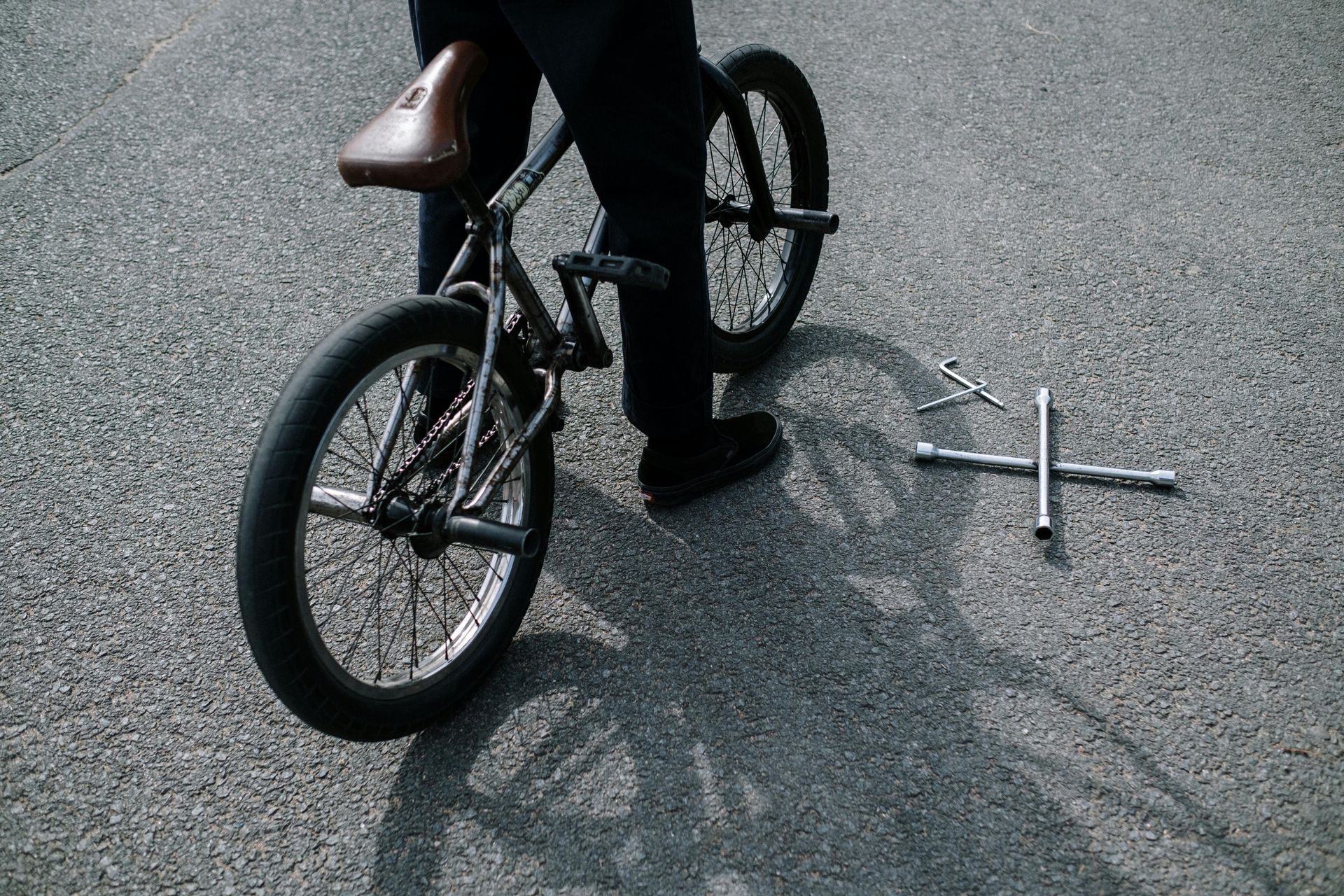 Person standing near a BMX bike, tool on ground, asphalt setting.