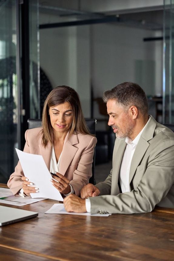 Woman and man in blazers reviewing paperwork at a wooden table in an office setting.