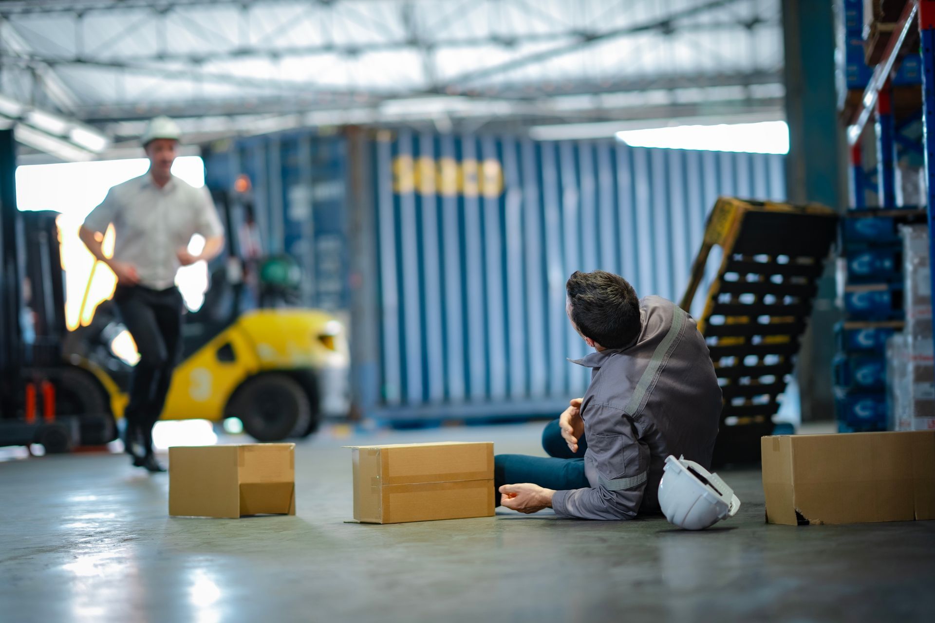 Warehouse scene with a worker seated on the floor near boxes while a forklift approaches in the background
