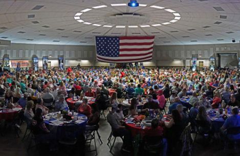 A large group of people are sitting at tables in a large room with an american flag hanging from the ceiling.