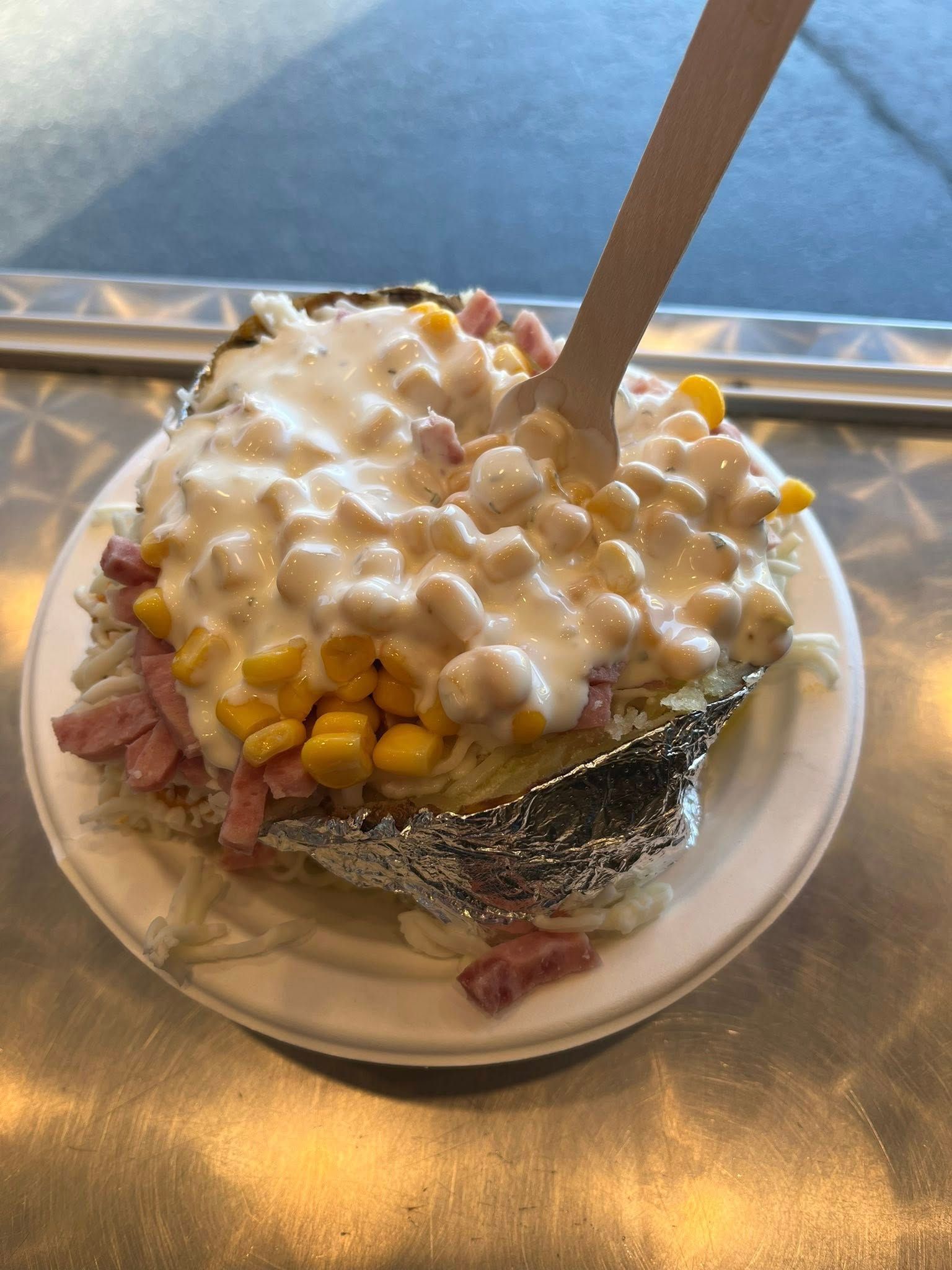 Baked potato with toppings, including corn and a creamy sauce, on a paper plate with a wooden fork.