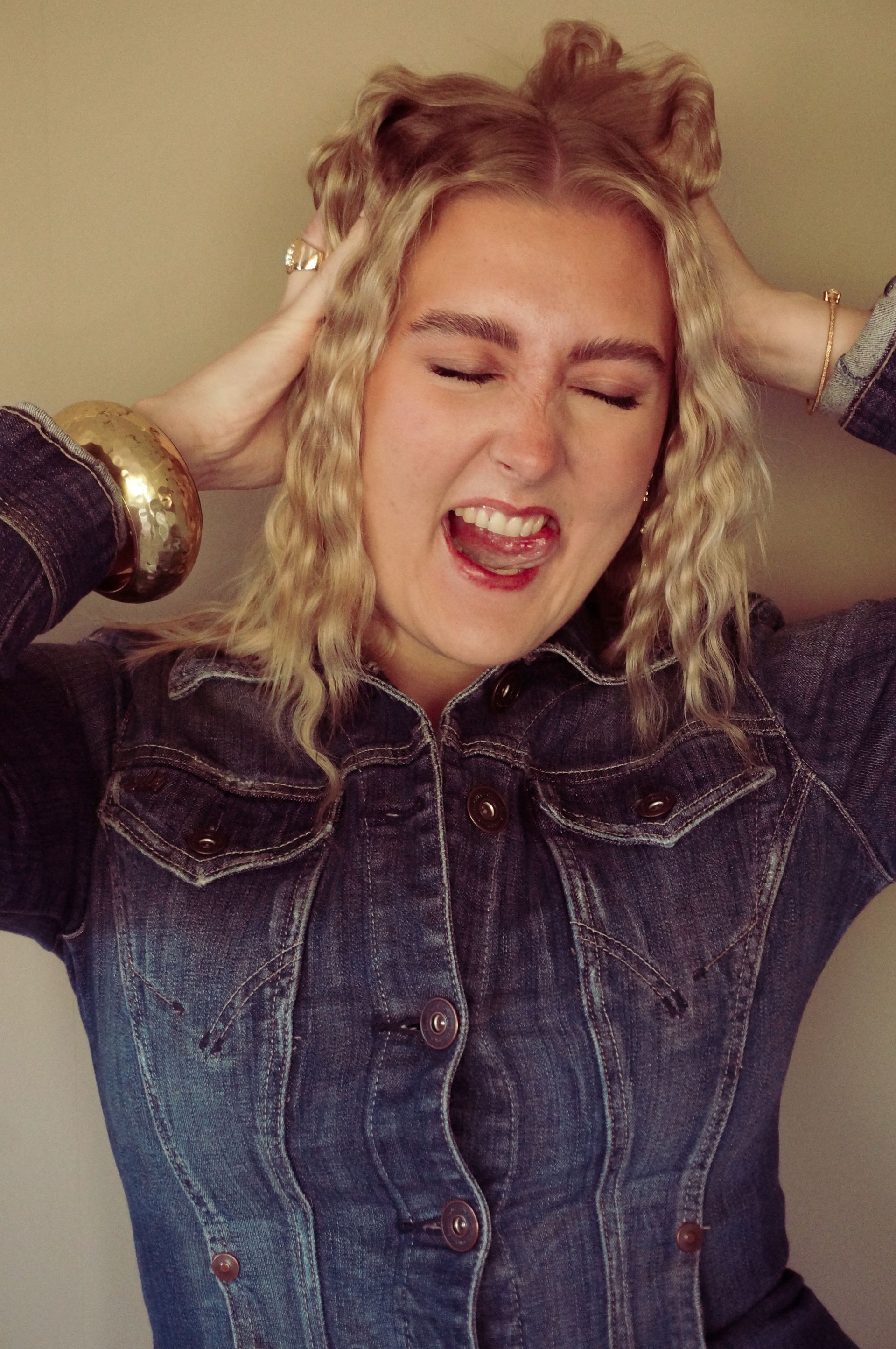 Woman with curly hair, eyes closed, mouth open in an expression of frustration, holding her head. Wearing a denim jacket.