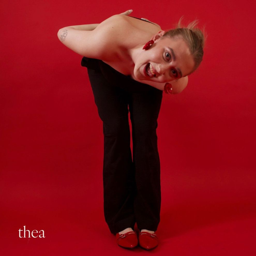 Person in black pants and red shoes, bends forward, arms crossed, mouth open, red background.